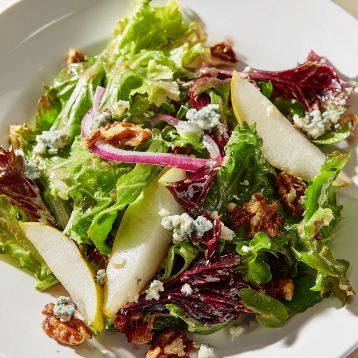 Fresh mixed winter greens topped with sweet sliced pears, crumbled blue cheese, and candied walnuts glistening in a light vinaigrette. Winter Green Salad with Pear and Candied Walnuts is plated elegantly on a white ceramic dish. Winter Green Salad with Pear and Candied Walnuts garnished with thinly sliced red onion and a drizzle of apple cider vinaigrette.