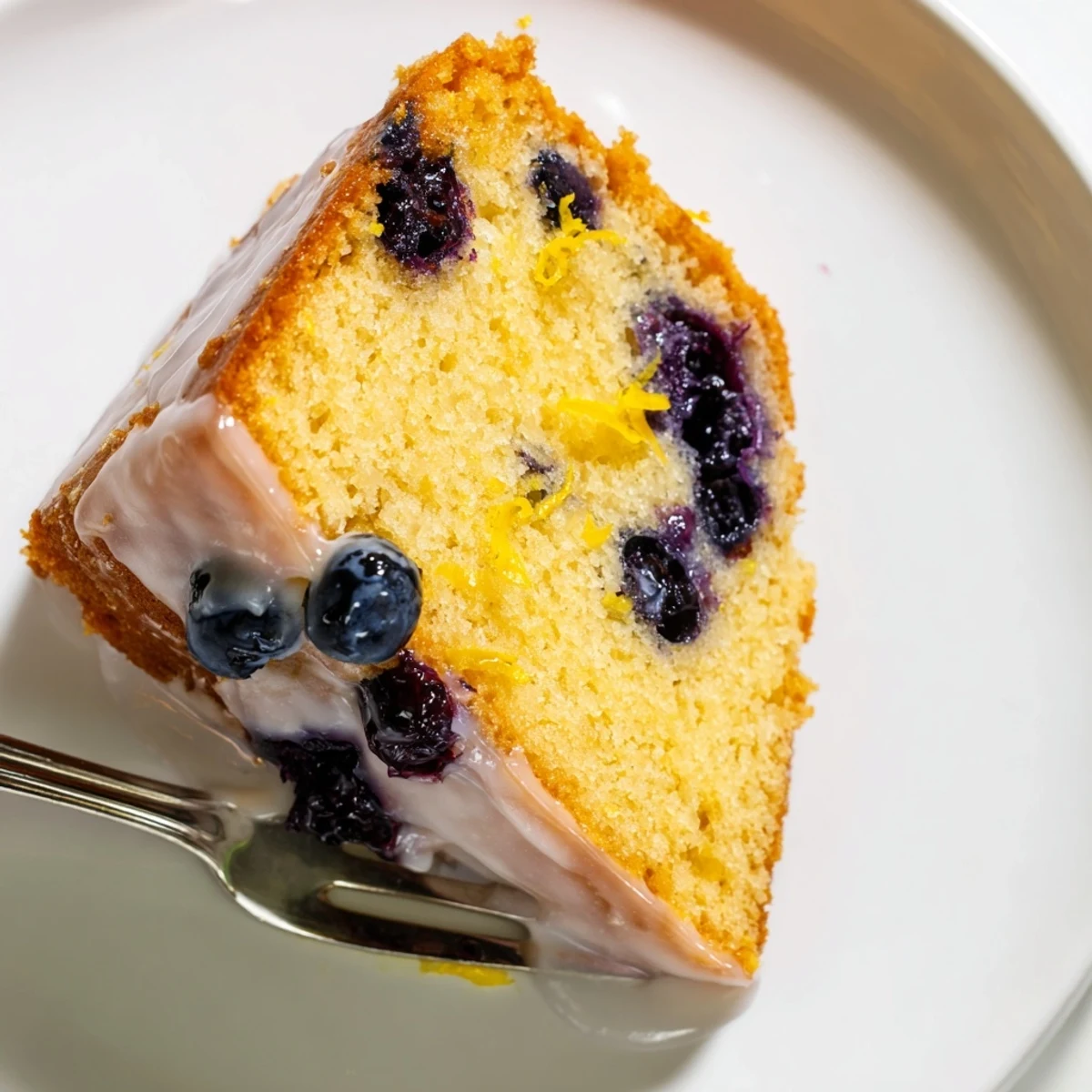 Moist lemon blueberry cake slice with glossy lemon glaze, fresh blueberries peeking from tender crumb, served on a white plate.
