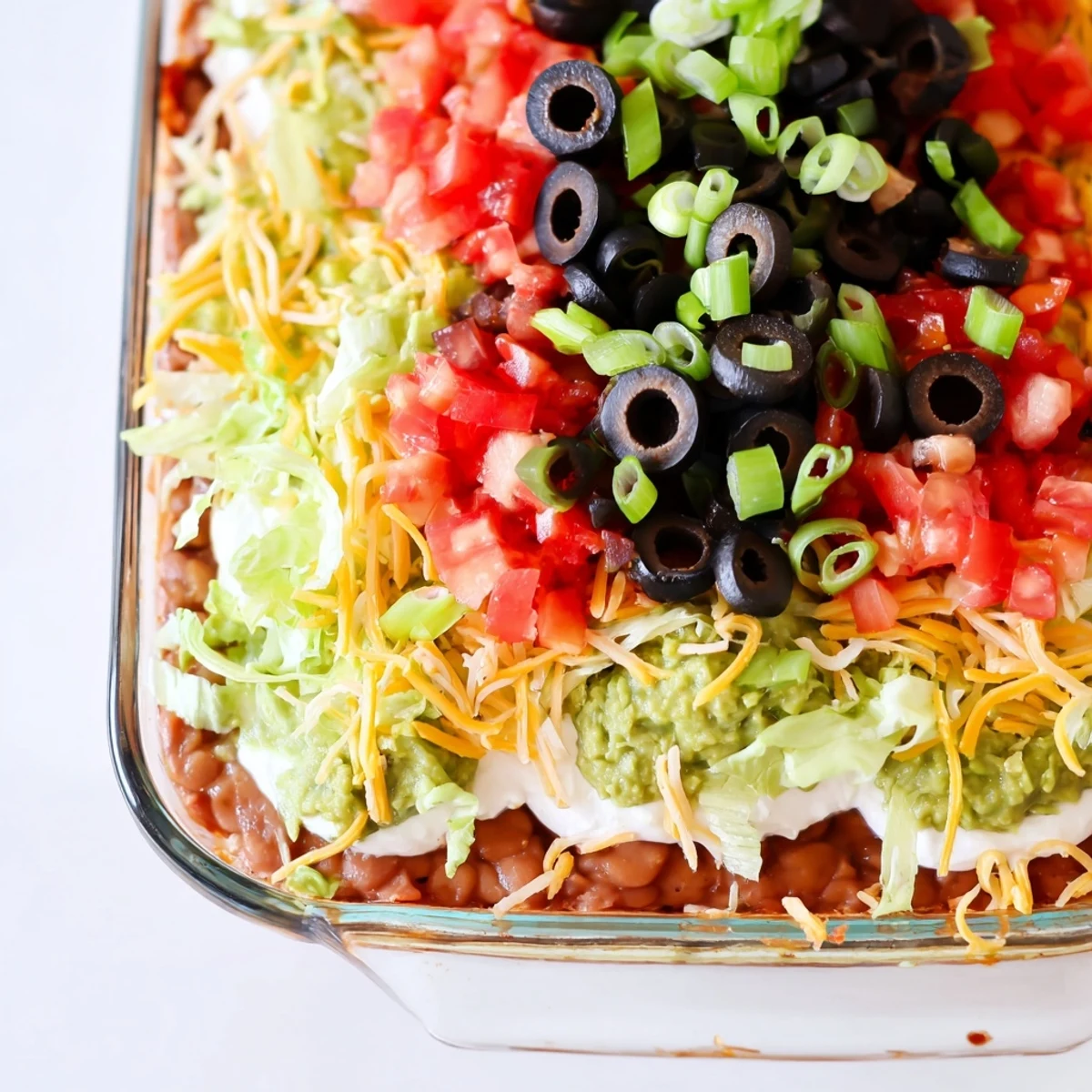 The Ultimate Seven Layer Taco Dip is garnished with fresh diced tomatoes and green onions, displayed in a glass baking dish for a party appetizer.