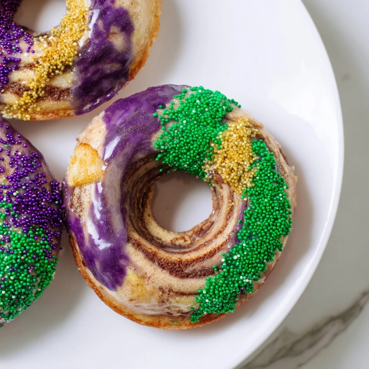 A close-up of a warm Mardi Gras King Cake Donut shows cinnamon swirls inside and a drizzle of sweet vanilla glaze.