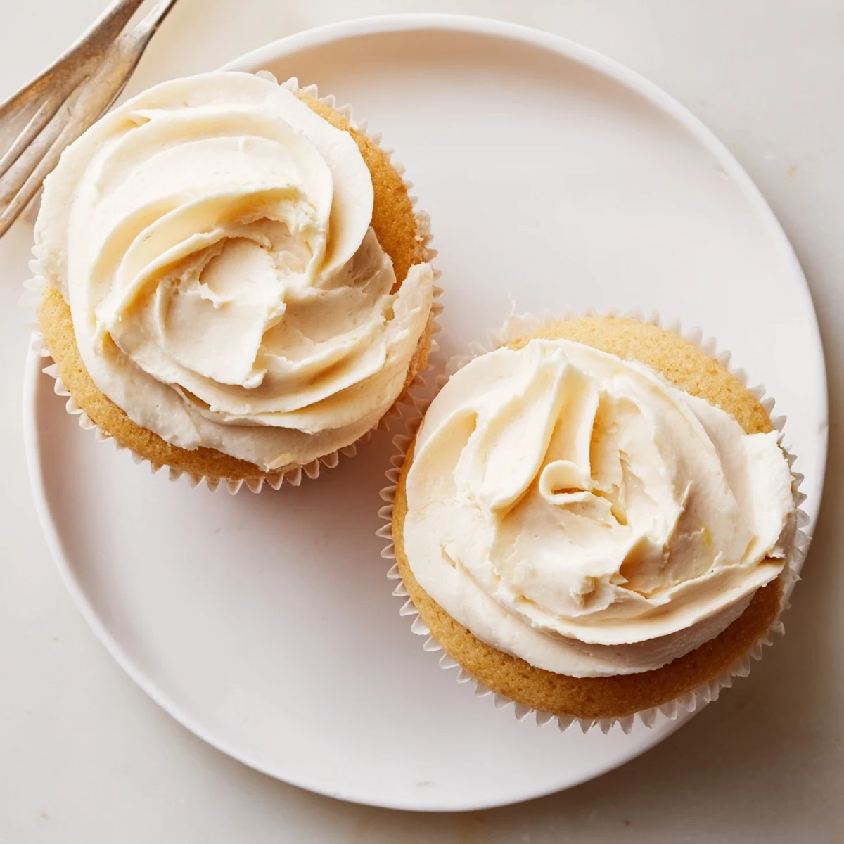 Newly frosted Elderflower Cupcakes sit on a wire cooling rack, surrounded by a small bowl of syrup and fresh lemon zest for a bright garnish idea.